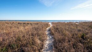 Ein sandiger Pfad führt durch ein trockenes, grasbewachsenes Gelände zum Strand.