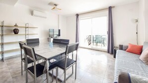 Un salon moderne avec une table à manger en verre, des chaises noires, un canapé, un téléviseur et un balcon aménagé avec du mobilier d’extérieur.