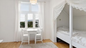 Een slaapkamer met een wit bed, een raam met gordijnen, een bureau met stoel en uitzicht op de omgeving.