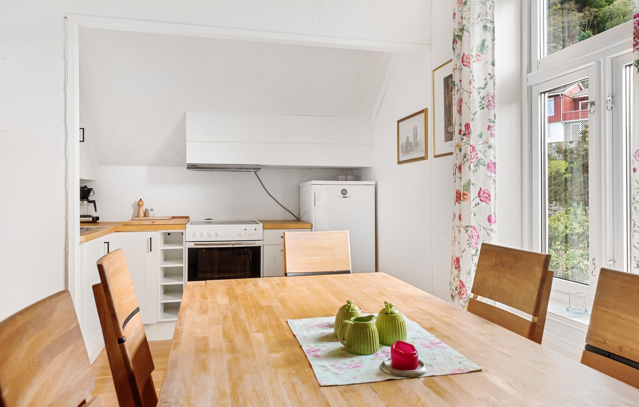Une cuisine compacte équipée d’une cuisinière, d’un four et d’un réfrigérateur. On y trouve une table à manger avec des chaises, ainsi qu’une fenêtre agrémentée de rideaux à motifs floraux.