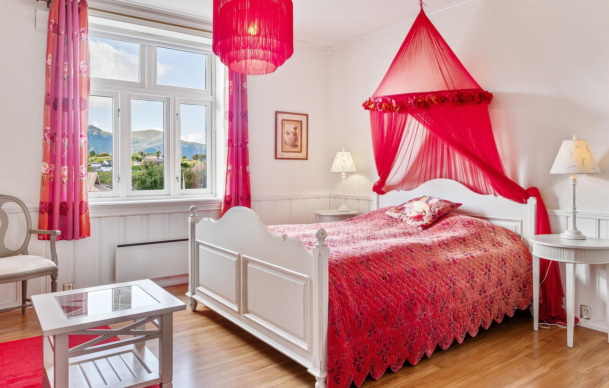 A bedroom with a white bed, red canopy, and a view of the outdoors through a window.