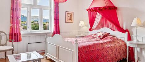 A bedroom with a white bed, red canopy, and a view of the outdoors through a window.