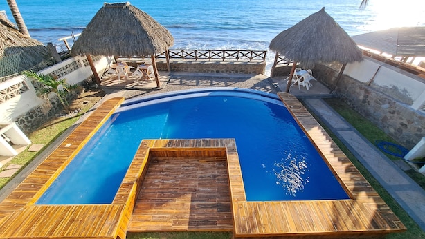 Ein Pool mit Holzterrasse, strohgedeckten Gebäuden und Blick auf das Meer.