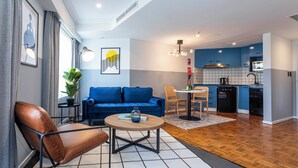 A modern living room with a blue sofa, a brown leather chair, and a round coffee table. A kitchen with blue cabinets and a black refrigerator is visible in the background. - GIA Sydney  (Sydney)