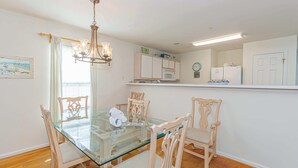 Uma sala de jantar com mesa de vidro e cadeiras de madeira, um lustre, uma cozinha com armários brancos e um relógio na parede.