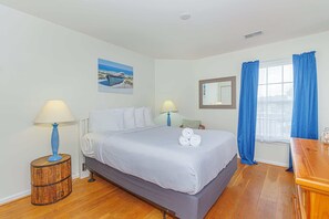 A bedroom with a bed, two bedside lamps, a window with curtains, a wooden side table, and a painting on the wall.