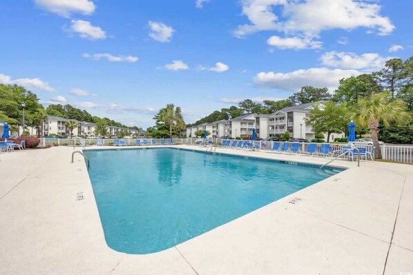 Una piscina rectangular con agua azul, rodeada por una terraza blanca. Se observan sillas reclinables azules y una valla blanca.