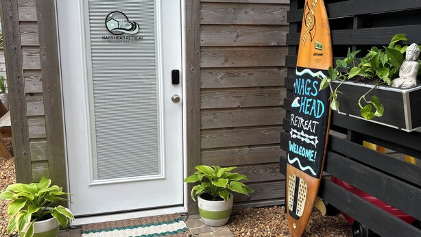 Uma porta de madeira com puxador de metal, um tapete de boas-vindas, placa com um prancha de surf e plantas em vasos.