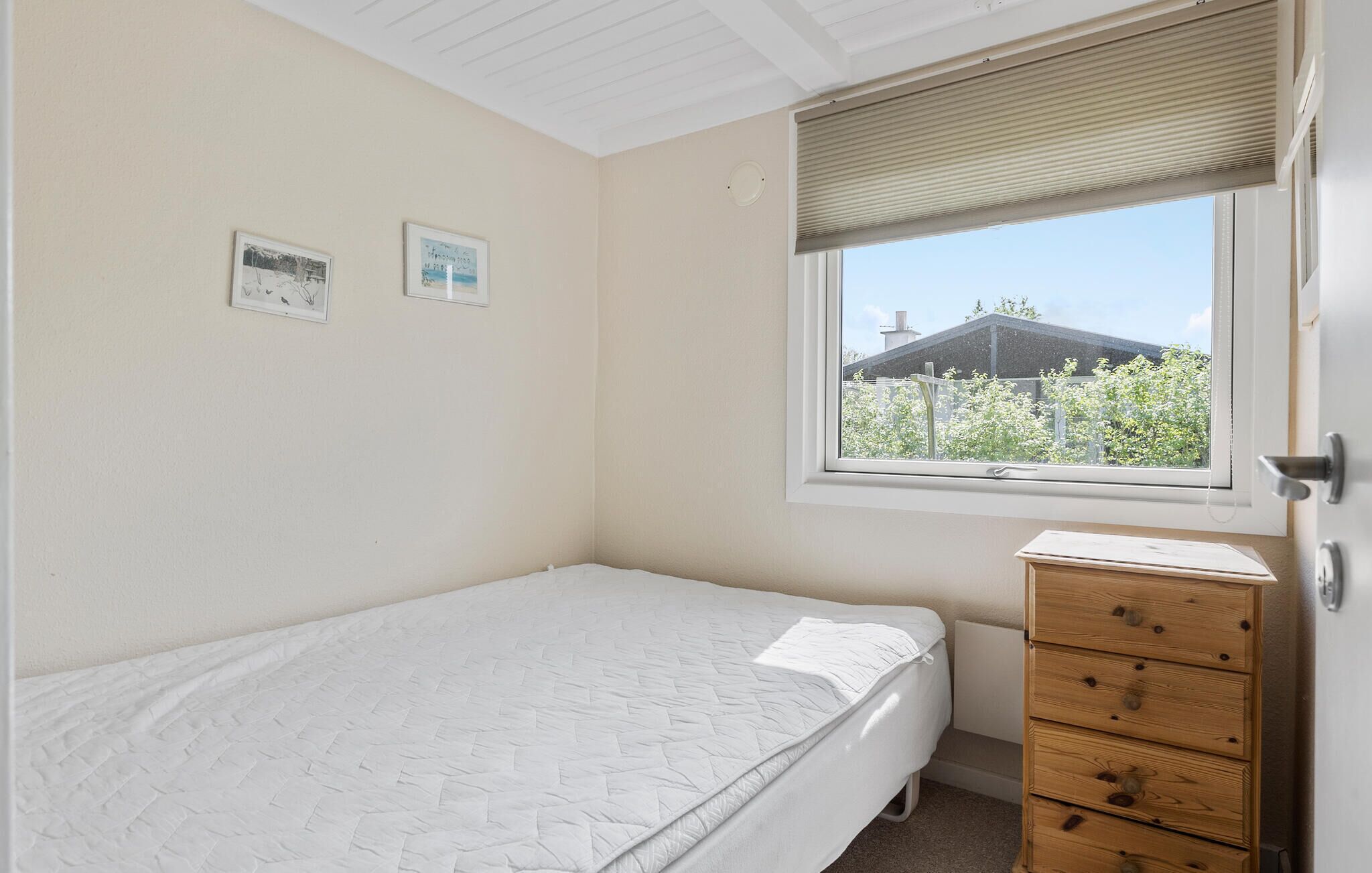 Een compacte, verzorgde slaapkamer met een eenpersoonsbed, een dressoir en een raam dat uitkijkt op groen en een gebouw.