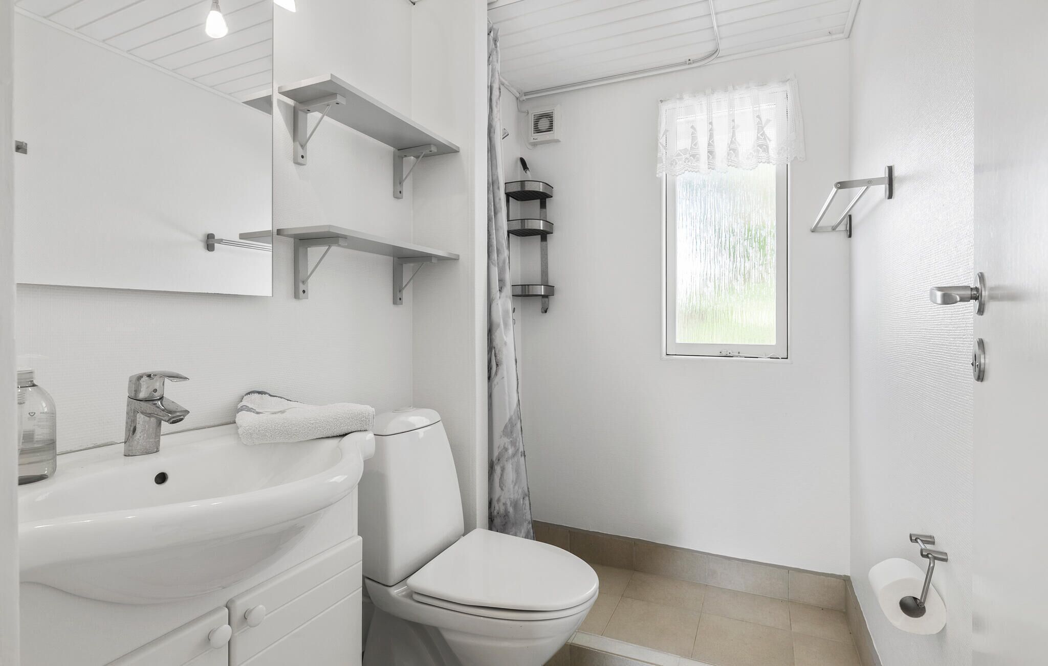 Ein modernes Badezimmer mit einem weißen Waschbecken, einer Toilette und einer Dusche. Es gibt Metallhandtuchhalter und ein Fenster mit Vorhang.