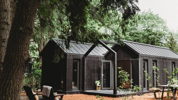 A modern cabin with a dark exterior, large windows, and a covered outdoor seating area with chairs and a table. - Hytte, which is Norwegian for cabin or (wooden) house in nature. (Epe)
