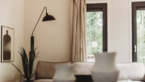 A living room with a sofa, a floor lamp, a plant, and a window with curtains. - Mysa Tiny House. At MYSA, every day feels as relaxed as a Sunday morning. (Epe)