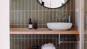 Een badkamer met een ronde spiegel, een witte wastafel, een houten kruk en een hangplant.