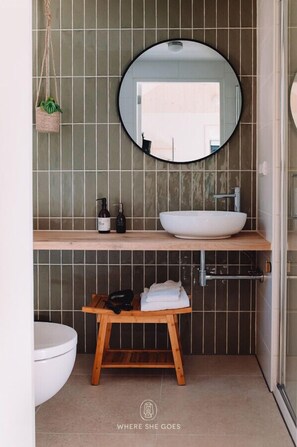 Une salle de bain avec un miroir rond, un lavabo blanc, un tabouret en bois et une plante suspendue.