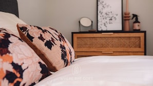 A neatly made bed with floral-patterned pillows, a bedside table with a framed picture, a round mirror, and a small wooden cabinet. - GrØn Tiny House in the forest (Epe)