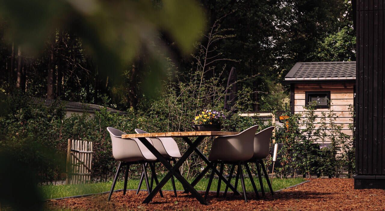 Een terras met een houten tafel en vier stoelen, omgeven door groen en een gebouw op de achtergrond.