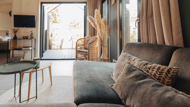 Una sala de estar acogedora con un sofá gris, una mesita redonda pequeña y un sillón de mimbre. Se ve una ventana grande con cortinas, y un televisor montado en la pared.