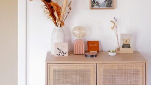 Quarto com parede branca, teto de madeira, um vaso branco com plantas secas, um abajur rosa e quadros emoldurados.