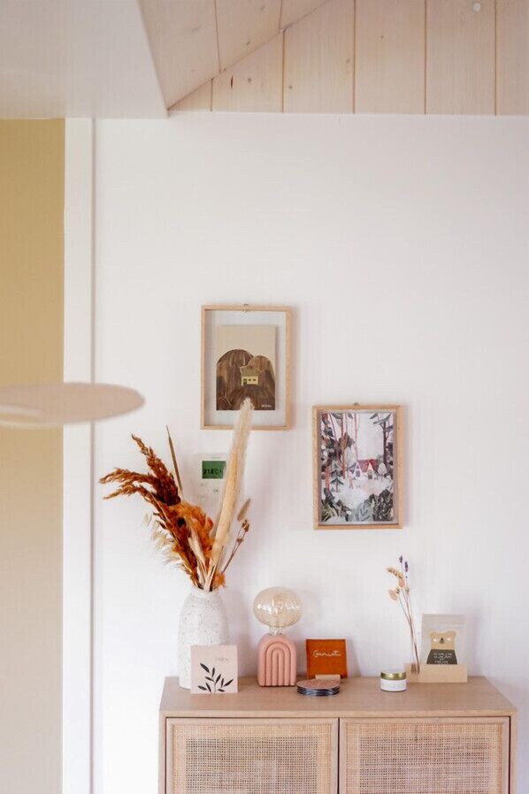 Una habitación con pared blanca, techo de madera, un jarrón blanco con plantas secas, una lámpara rosa y cuadros enmarcados.