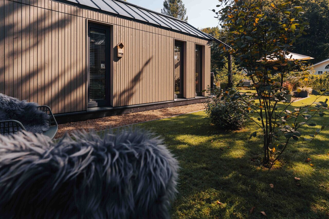 A modern wooden building with a metal roof, a small garden, and a visible door.