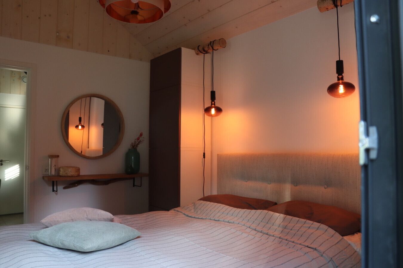 A bedroom with a bed, a round mirror, a shelf with decorative items, and two hanging light fixtures.