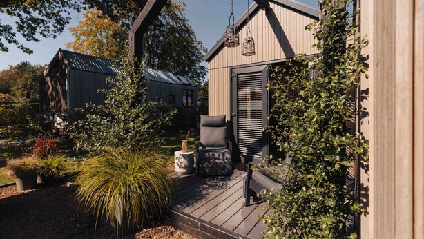 Une terrasse en bois avec un banc et des plantes en pot, un luminaire suspendu et un bâtiment doté d’un toit en pente et de volets.
