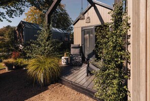 Eine Holzterrasse mit einer Bank und Topfpflanzen, einer Hängelampe sowie einem Gebäude mit schrägem Dach und Fensterläden.