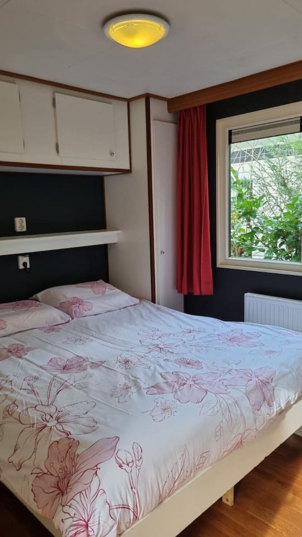 A bedroom with a bed, a window with a view of greenery, a radiator, and a ceiling light.