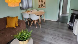 Uma sala de estar moderna com um sofá marrom, uma mesa de centro redonda com uma planta, uma área de jantar com uma mesa de madeira e cadeiras brancas, e uma cozinha com um cesto de lixo preto e um eletrodoméstico branco.