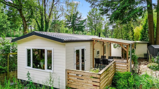 A small, single-story cabin with a porch and a deck, surrounded by dense forest.
