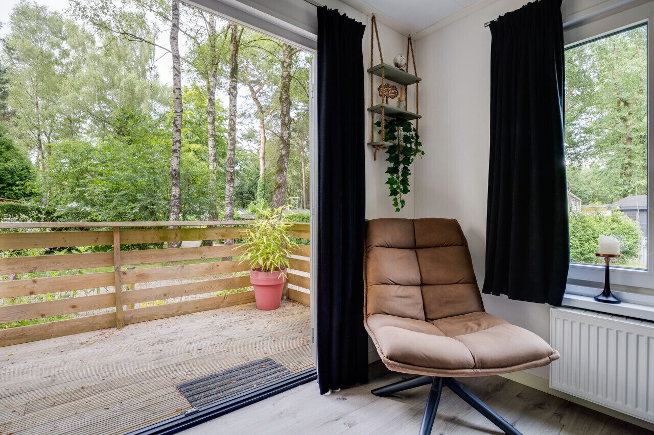 A balcony with a wooden railing, a potted plant, a cushioned chair, and a window with black curtains.