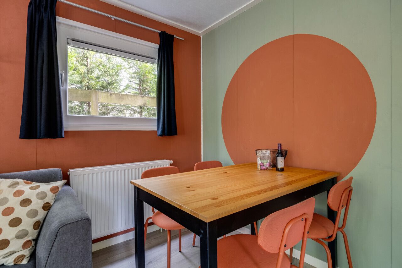 Un comedor con mesa y sillas de madera, un radiador, una ventana con cortinas y una gran decoración naranja en la pared.