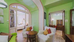 Une pièce avec un mur vert, un canapé blanc, une table en bois et un lit agrémenté d’un rideau à motifs floraux.