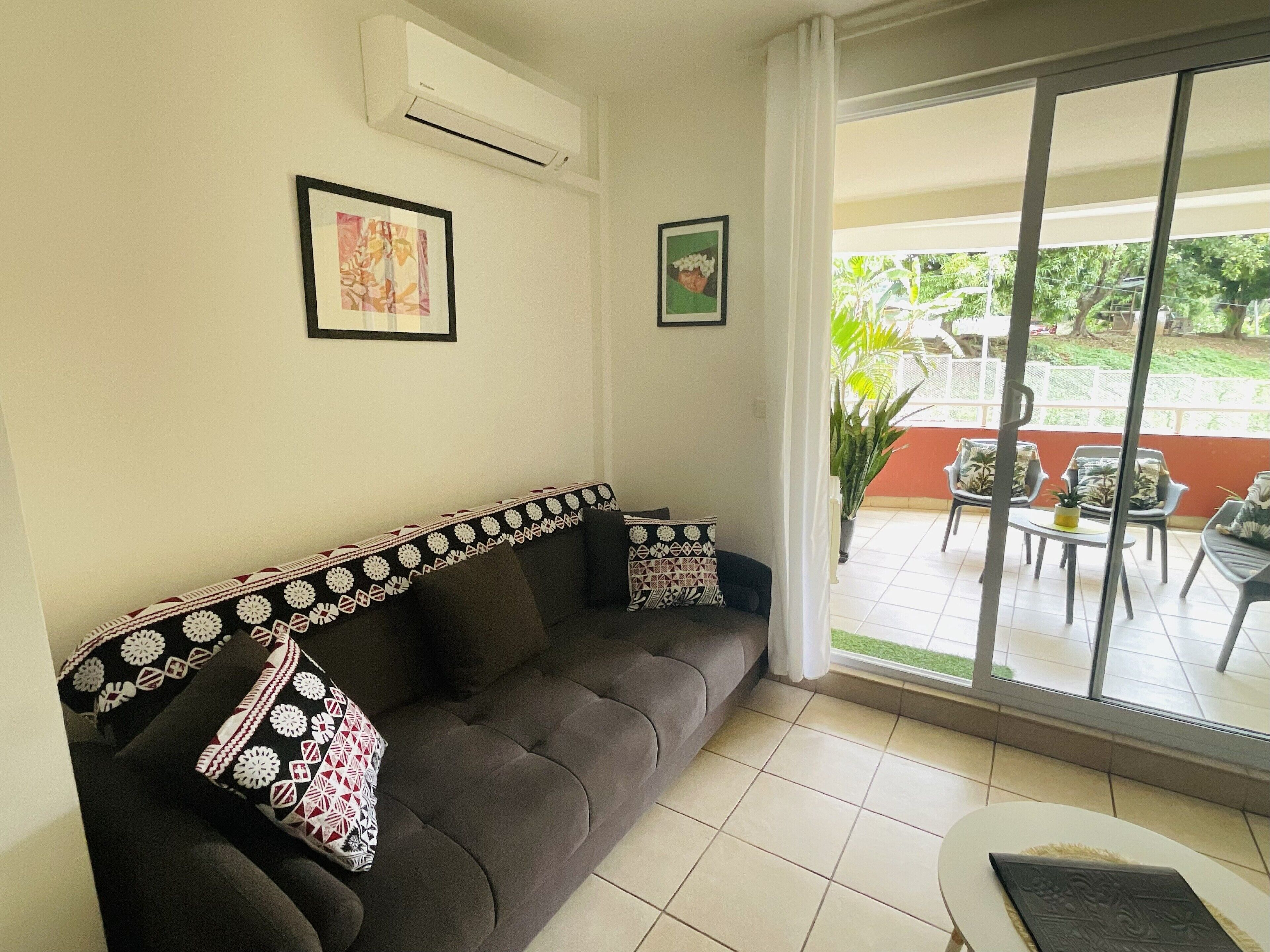 A living room with a dark brown sofa, two patterned pillows, and a small round table. There are two framed pictures on the wall, and an air conditioning unit above the sofa. A sliding glass door leads to an outdoor patio area with chairs and a table.