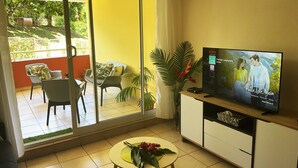 A living room with a television displaying a movie, a balcony with chairs and plants, and a small table with flowers. - Prestige Lodging Papeete (Papeete)