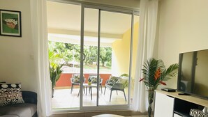 A living room with a sofa, a coffee table, a TV, and a sliding glass door leading to an outdoor patio with chairs and plants. - Prestige Lodging Papeete (Papeete)