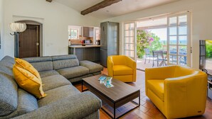 Una sala de estar con un sofá, sillones amarillos y una mesa de centro. Se ve una cocina al fondo con vistas al exterior.