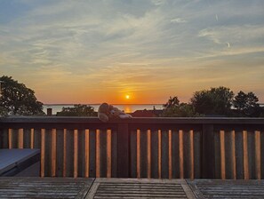 Une vue sur le coucher de soleil depuis une terrasse en bois dotée d’une balustrade, donnant sur une étendue d’eau et des arbres.