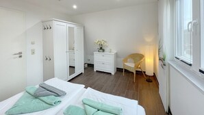 A modern bedroom with two beds, a wardrobe, a bedside table with a vase, a wicker chair, and a window with curtains. - Meeresbrise - Stilvoll Genießen, auf 80qm, für bis zu 4 Personen, auf der Sonneninsel Usedom (Loddin)