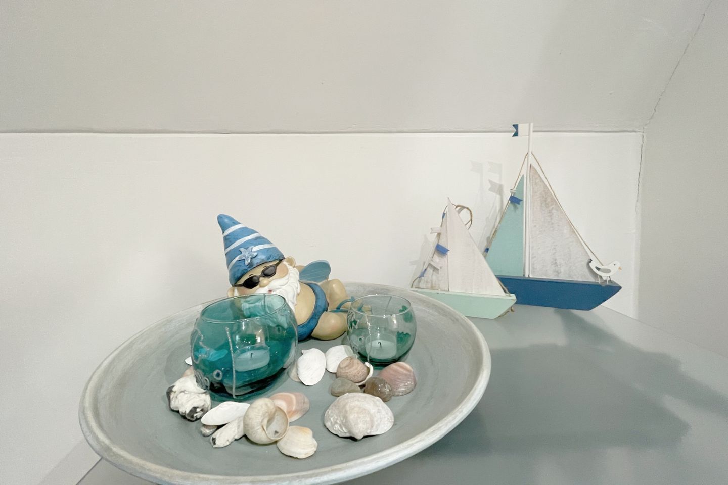 Escena decorativa con una figurita de gnomo, un velero y conchas marinas sobre un plato de cerámica.