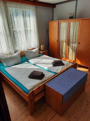 A wooden bed with white linens and a bench, a wardrobe with glass doors, and sheer curtains. - Bungalow Waldstrasse - Charmanter Ferienbungalow auf 55qm für 5 Personen (Ückeritz)