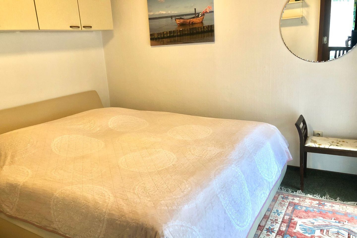 A neatly made bed with a patterned bedspread, a small wooden chair, a framed picture of a boat on the wall, and a round mirror.