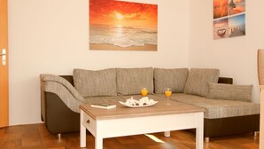 Sala de estar com sofá, mesa de centro com um livro e um prato de comida, e uma pintura de pôr do sol na parede.