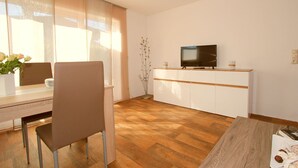 Quarto moderno e bem iluminado, com piso de madeira, mesa de jantar com cadeiras, televisão em um móvel branco e um vaso com flores.