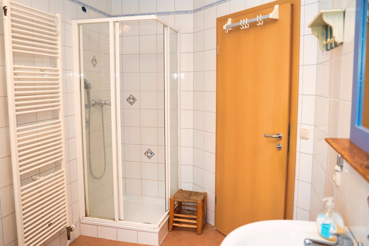 A bathroom with a glass-enclosed shower, a wooden door, a small wooden stool, and a white sink with a soap dispenser.