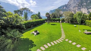 A well-maintained garden with a stone pathway leading to a seating area, surrounded by lush greenery and a view of a mountainous coastline.