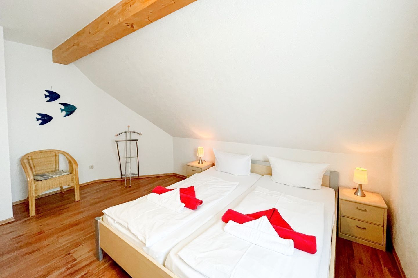 Quarto com teto inclinado, cama com roupa de cama branca e detalhes vermelhos, cadeira de madeira, mesa de cabeceira com abajur e arte na parede com tema de peixes.