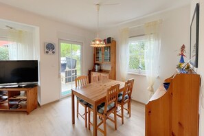 Uma área de refeições com uma mesa e cadeiras de madeira, uma televisão em um suporte, um armário com portas de vidro e uma janela com cortinas.