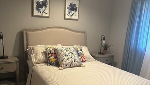 A neatly made bed with a quilted cover, two framed floral paintings on the wall, a bedside lamp, and a window with curtains. - Cozy apartment in Pendleton Oregon  (Pendleton)