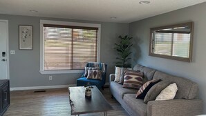 A living room with a blue armchair, a grey sofa, and a wooden coffee table. There are large windows with blinds, a potted plant, and a framed mirror on the wall. - Cozy apartment in Pendleton Oregon  (Pendleton)